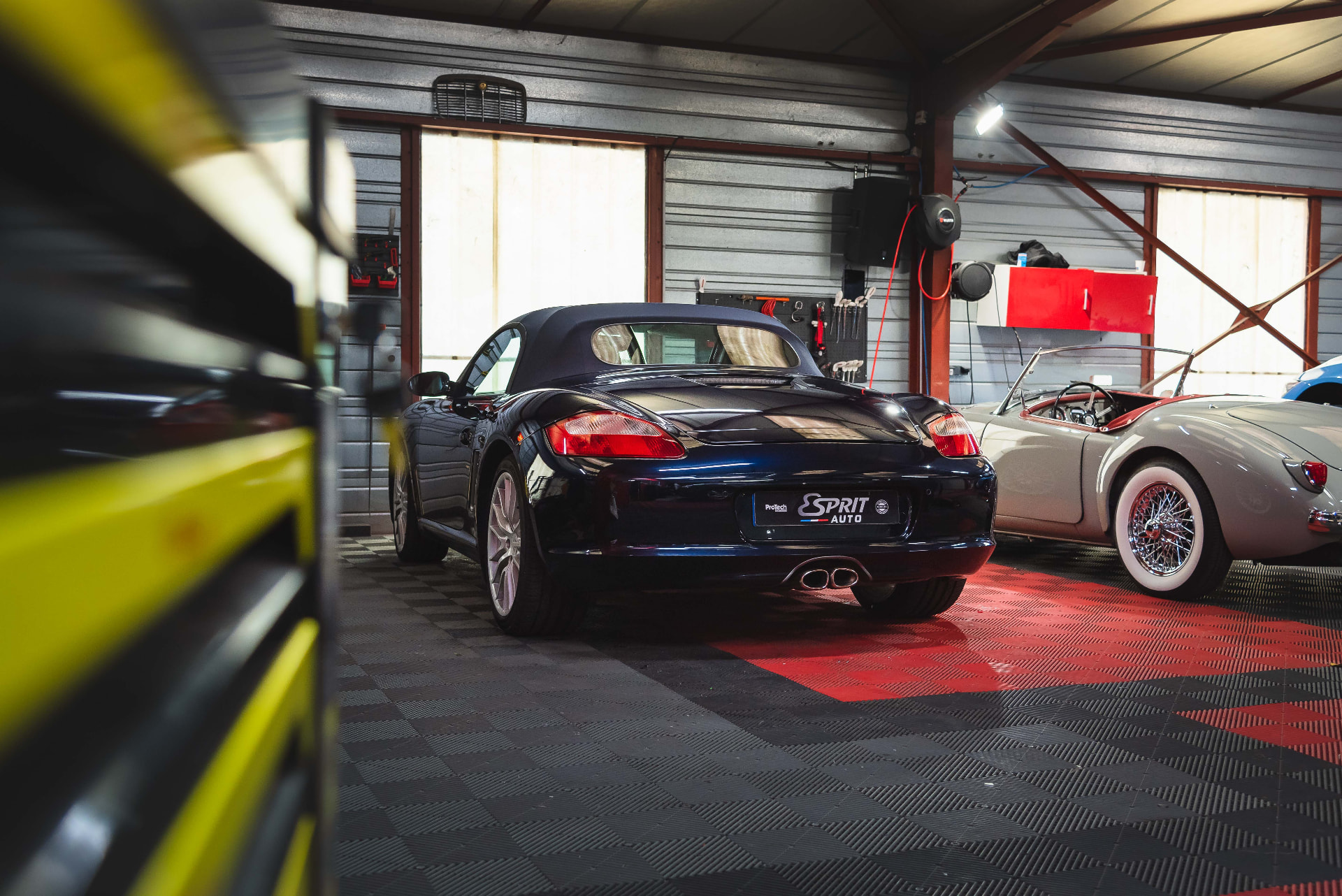 Porsche Boxster noire dans l'atelier Esprit Auto, entre une carrosserie floue et un roadster classique