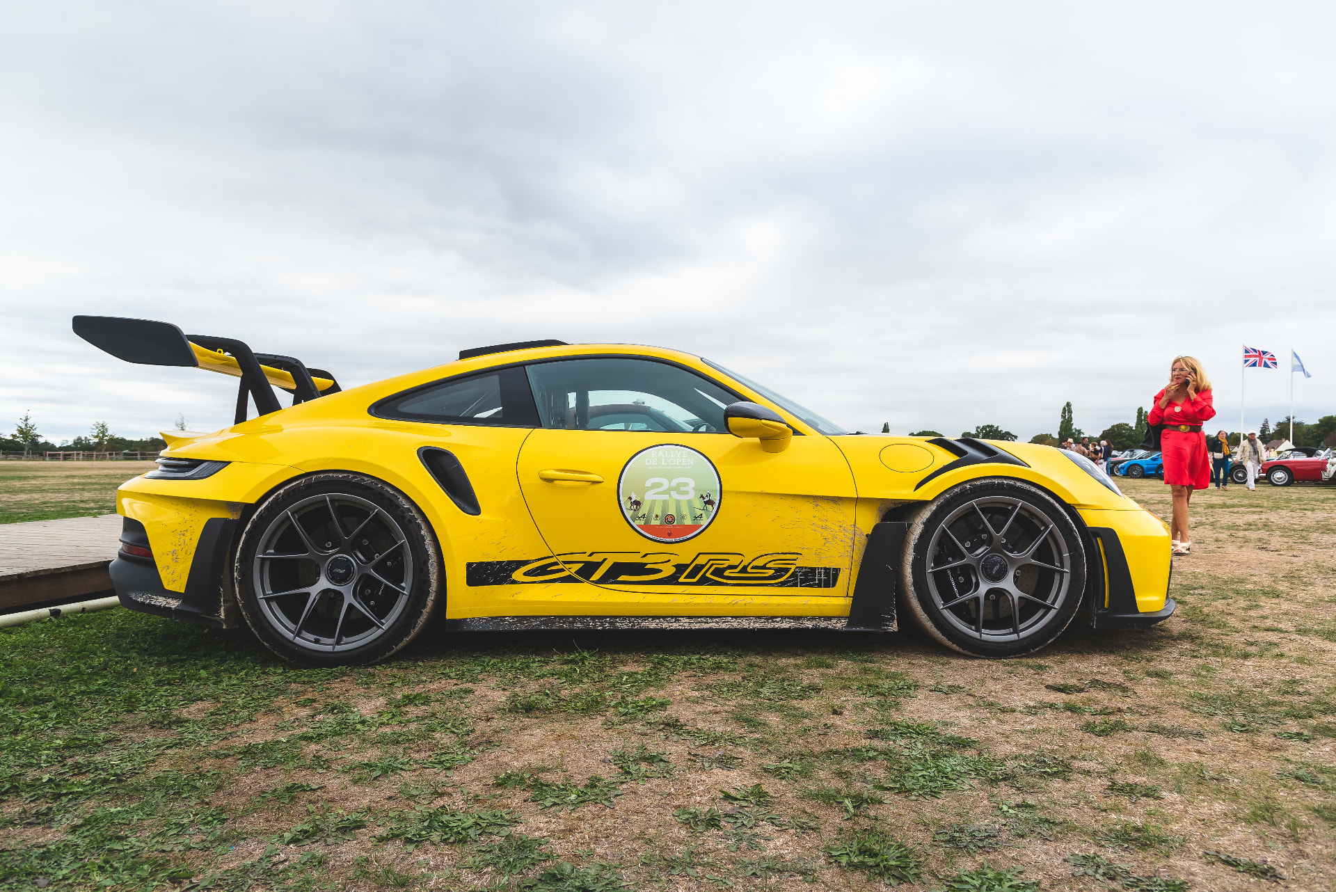 Profil d'une Porsche jaune au rallye, drapeaux et participants en arrière-plan