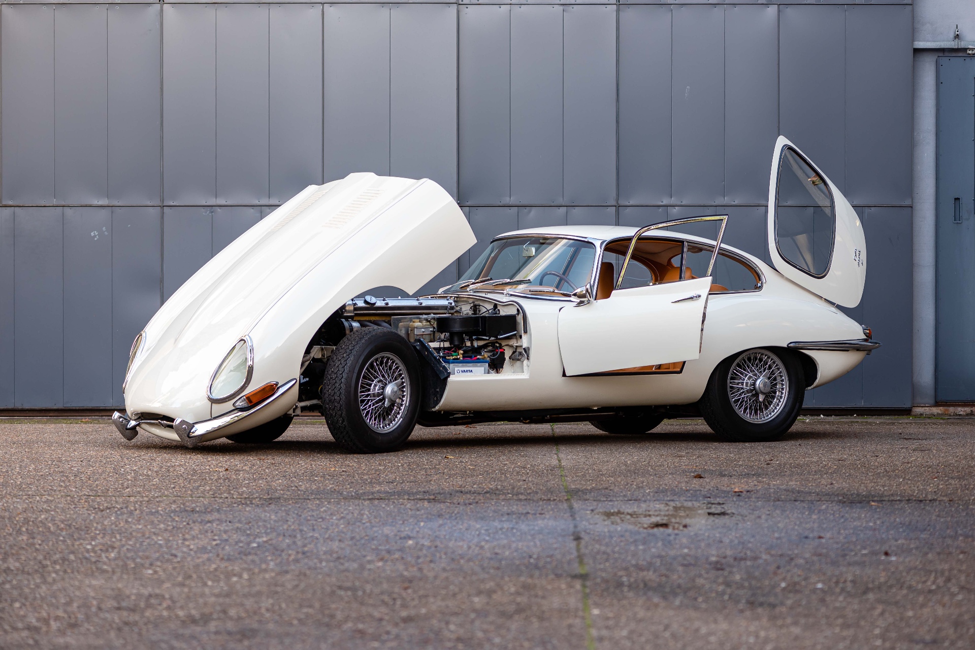 Jaguar Type E blanche, capot avant et hayon arrière ouverts devant un atelier
